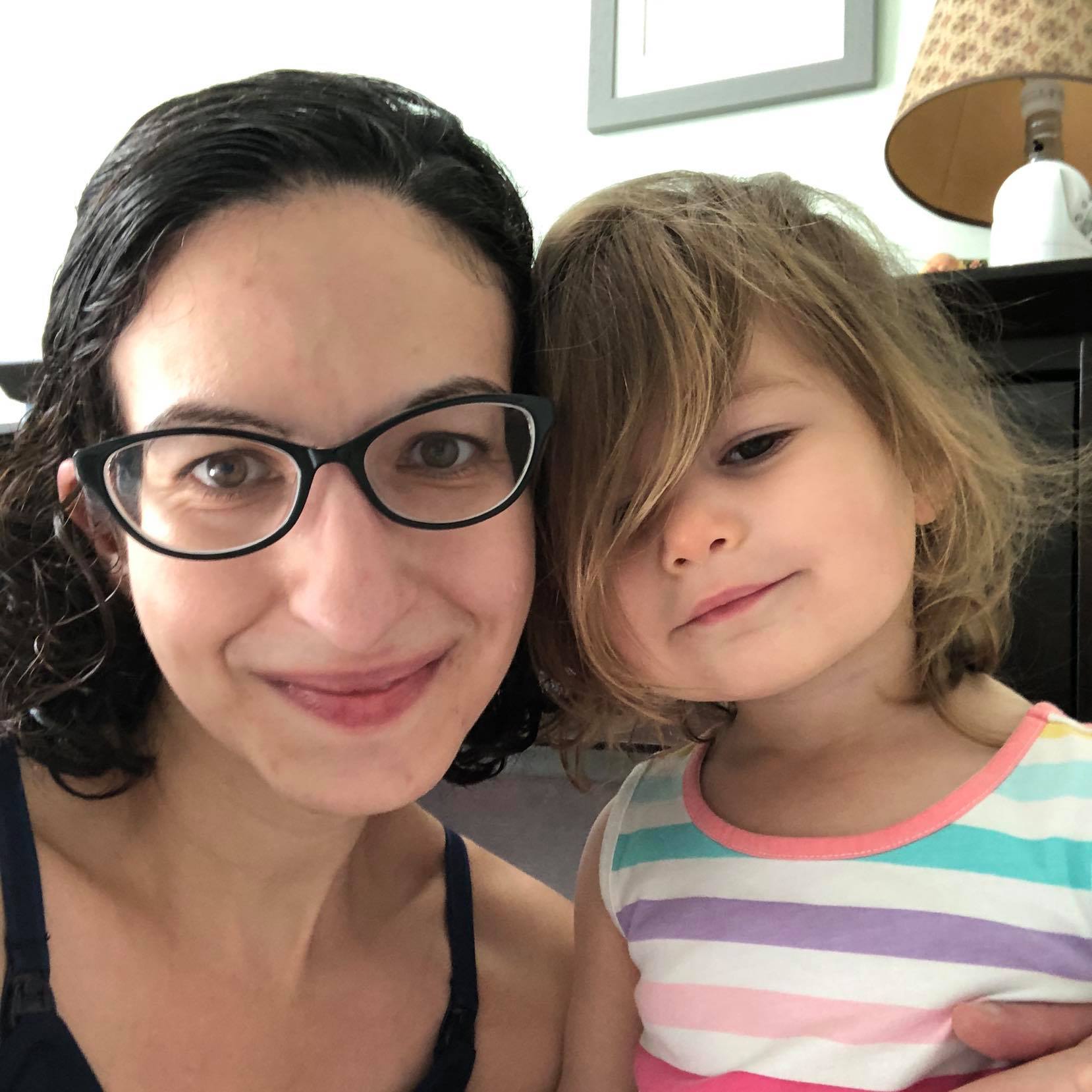 A close-up selfie of Joni, a white woman with dark curly hair and glasses, and her 2-year old daughter, a young white girl with short wavy dark blonde hair.