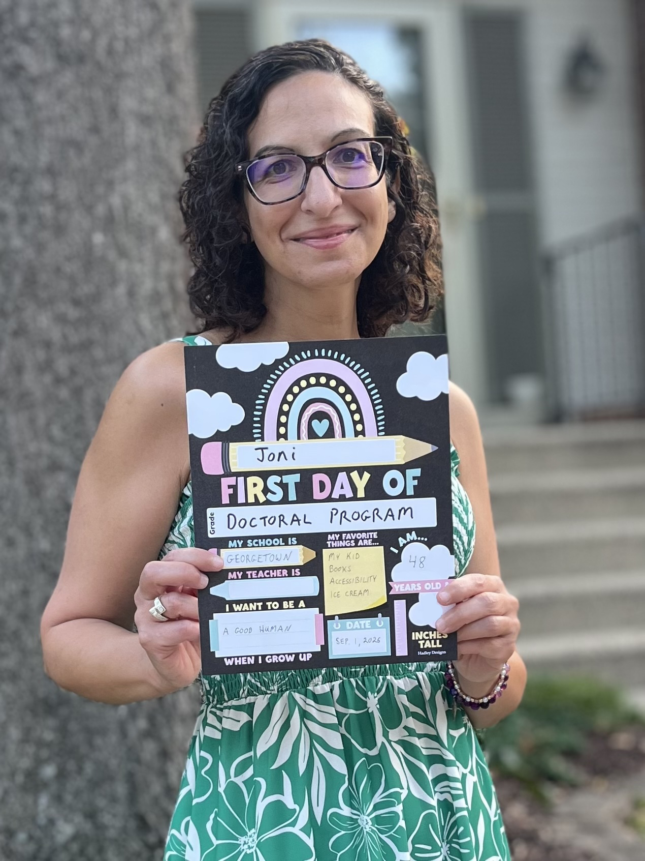 Joni, a light-skinned Arab woman with dark curly hair and glasses, stands outside holding a “first day of school” sign.