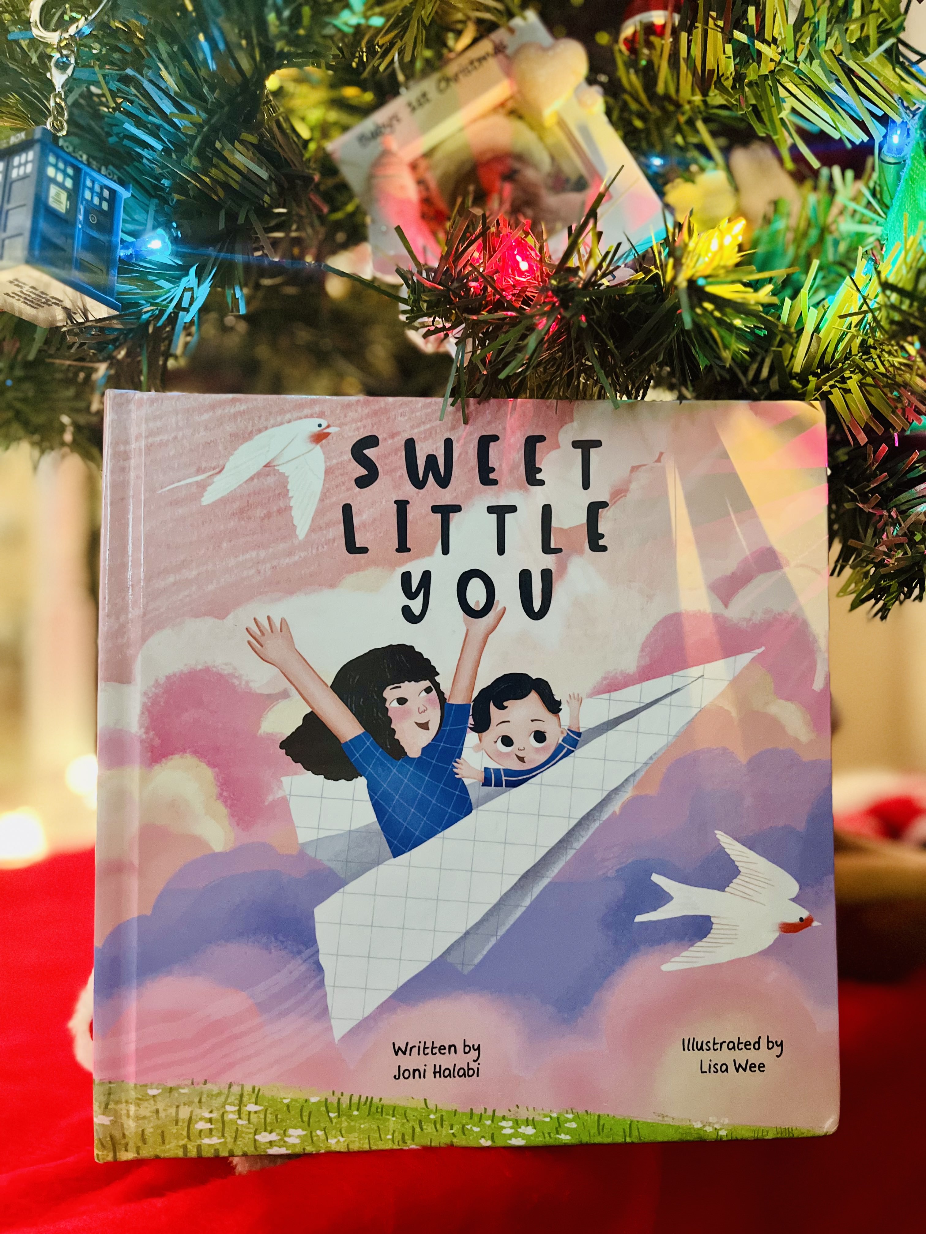 A hardcover copy of Sweet Little You is sitting under a holiday tree. The book cover features a mom and baby flying in a paper airplane